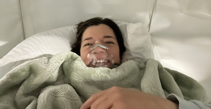 Coach Jenn Experiences HBOT smiling in a clear oxygen mask wrapped in blanket